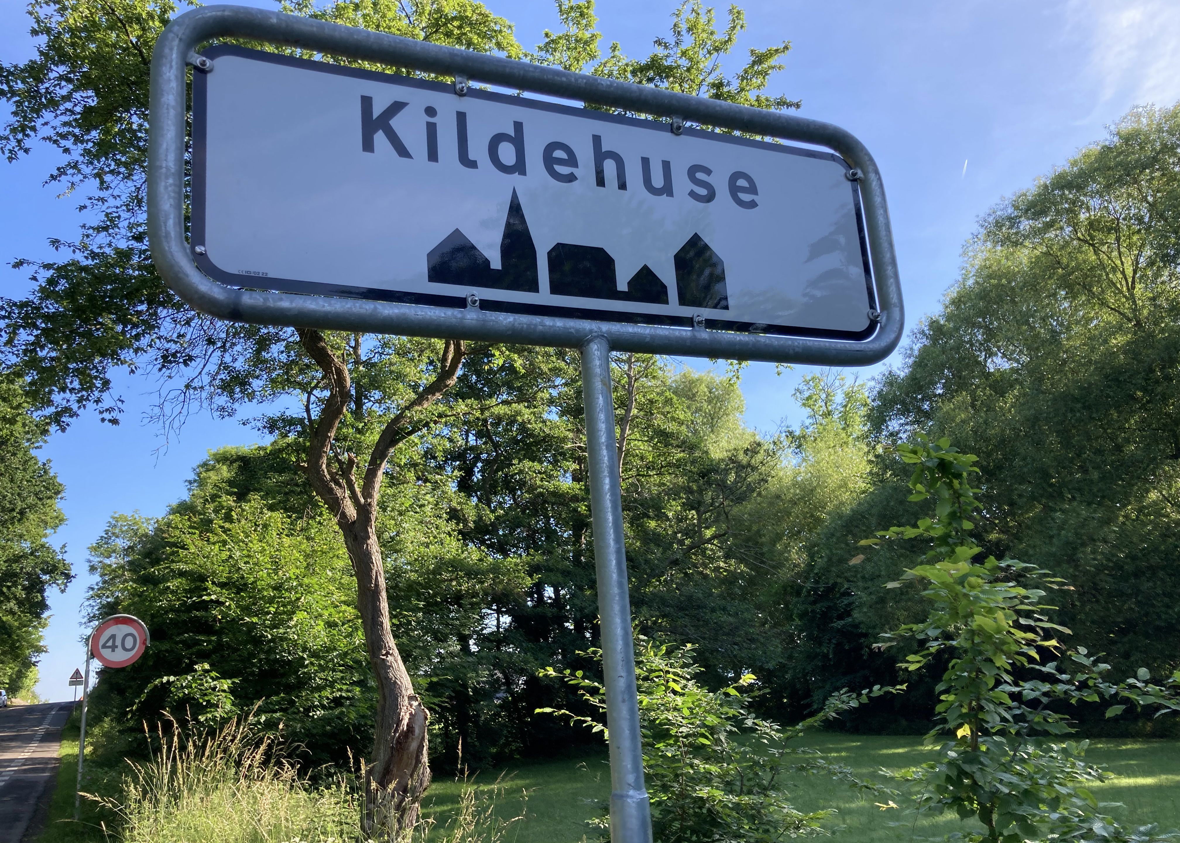 Vejskilt / byzonetavle med teksten "Kildehuse" med solbeskinnede træer i baggrunden