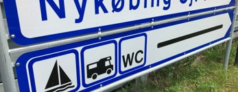 Blåt/hvidt vejskilt med teksten "Nykøbing Sjælland Havn" og ikoner for båd, autocamper og WC