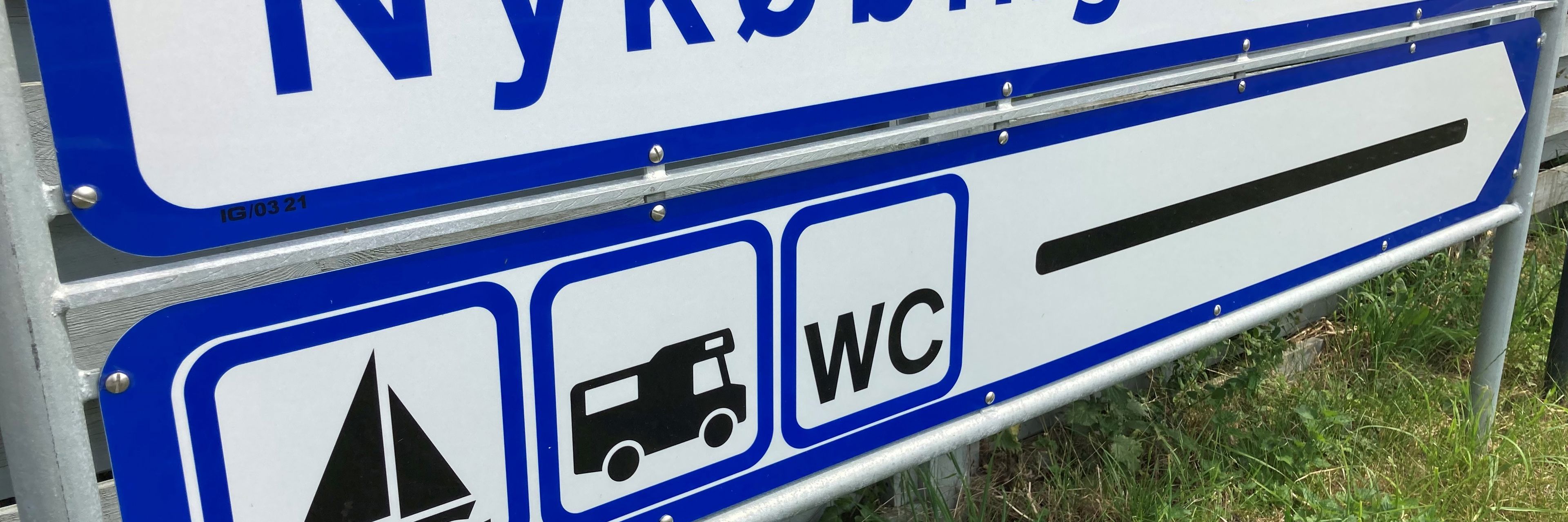 Blåt/hvidt vejskilt med teksten "Nykøbing Sjælland Havn" og ikoner for båd, autocamper og WC
