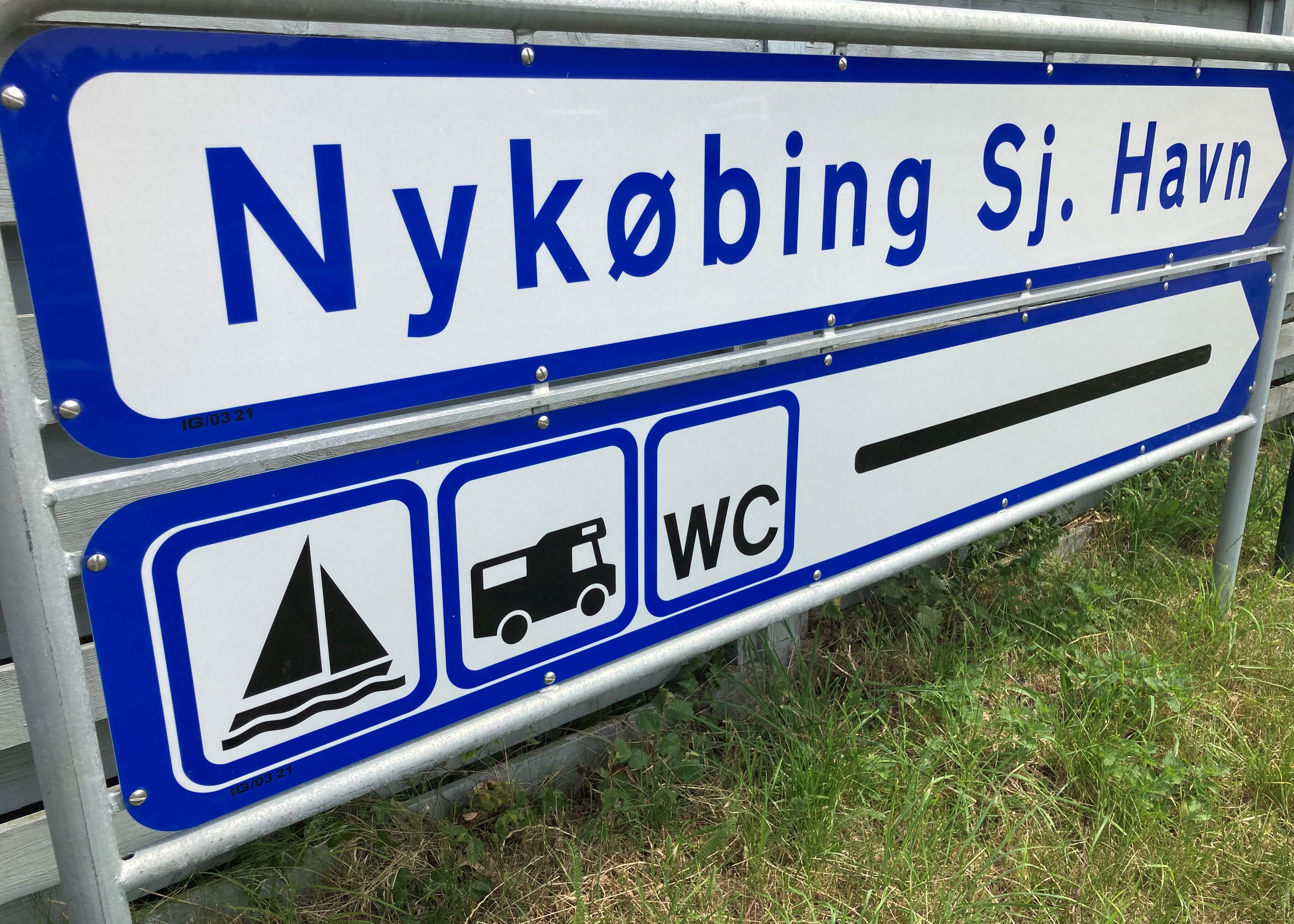 Blåt/hvidt vejskilt med teksten "Nykøbing Sjælland Havn" og ikoner for båd, autocamper og WC