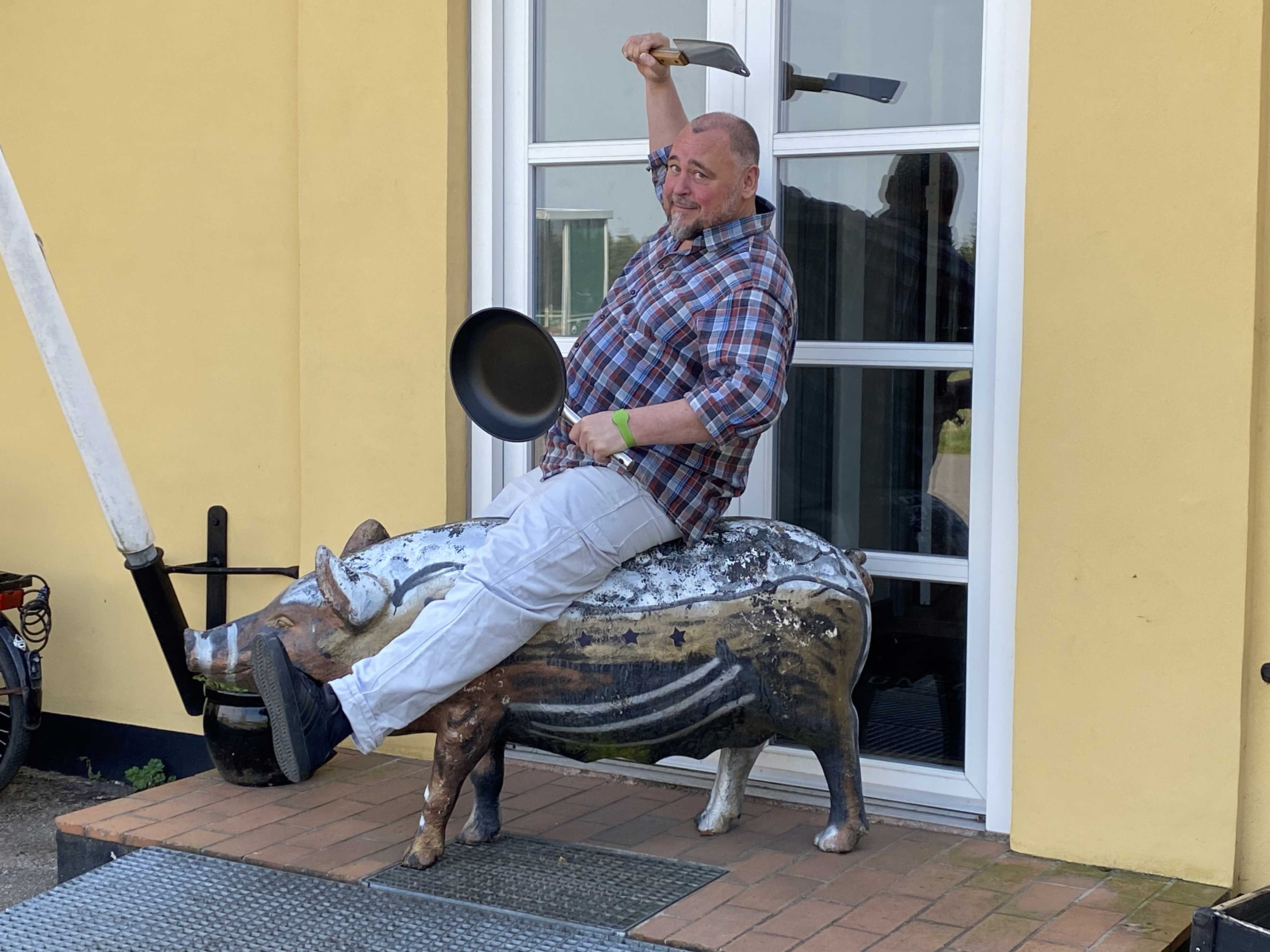 Mand der rider på en plastikgris med en stegepande og en kødkniv i hænderne