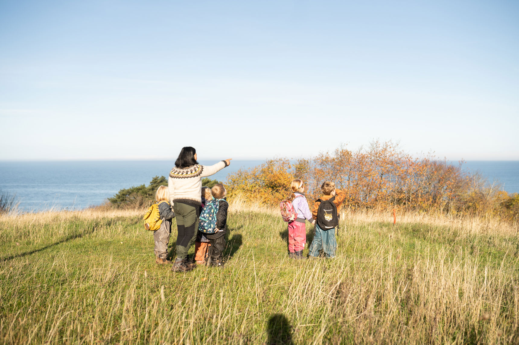 En voksen og flere børnehavebørn der står og ser ud over havet