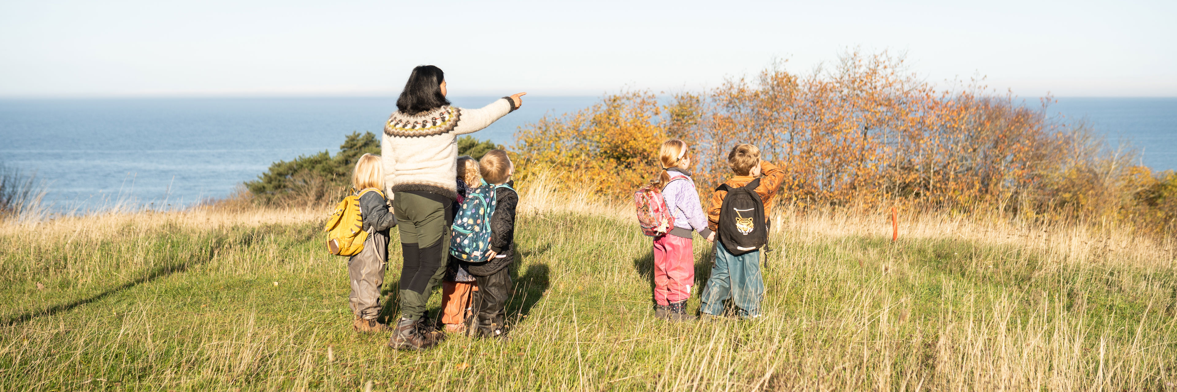 En voksen og flere børnehavebørn der står og ser ud over havet