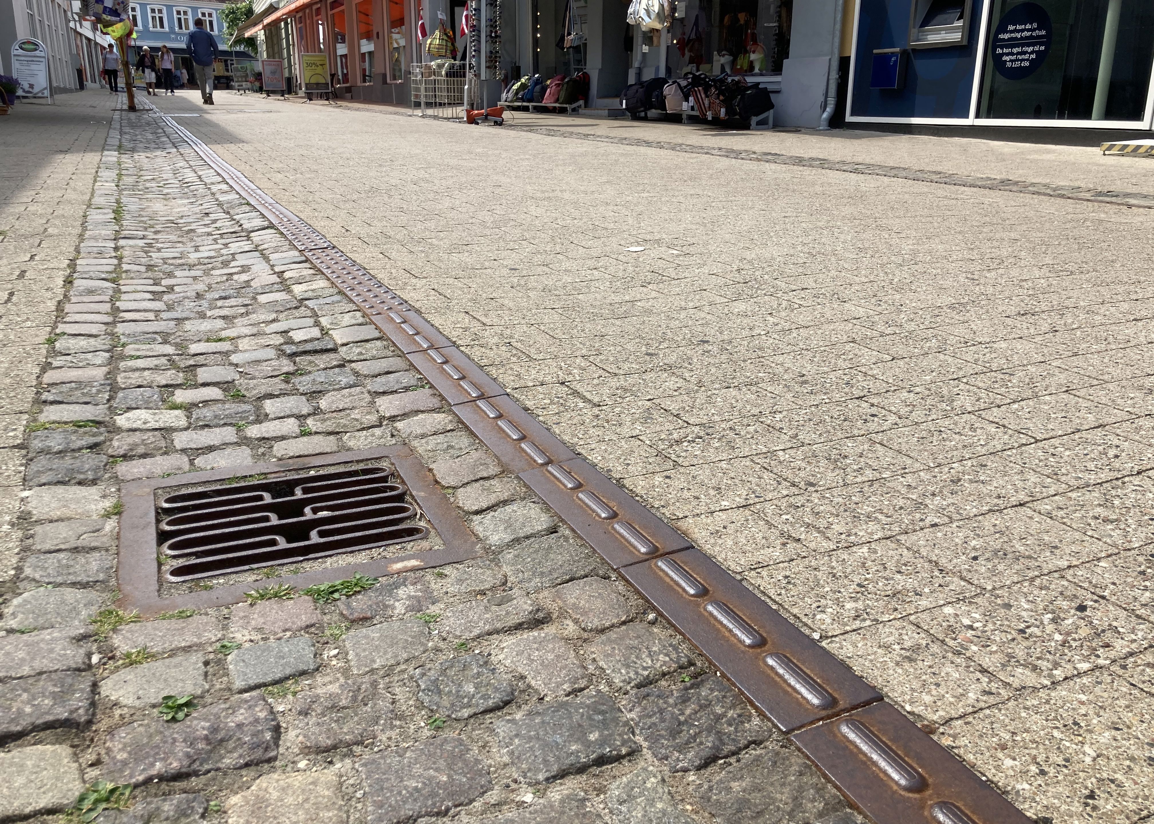 Closeup af brostensbelægningen i Algade, hvor gågaden er i fokus og fremstår nyfejet. I baggrunden fornemmer man handelslivet og butikkerne