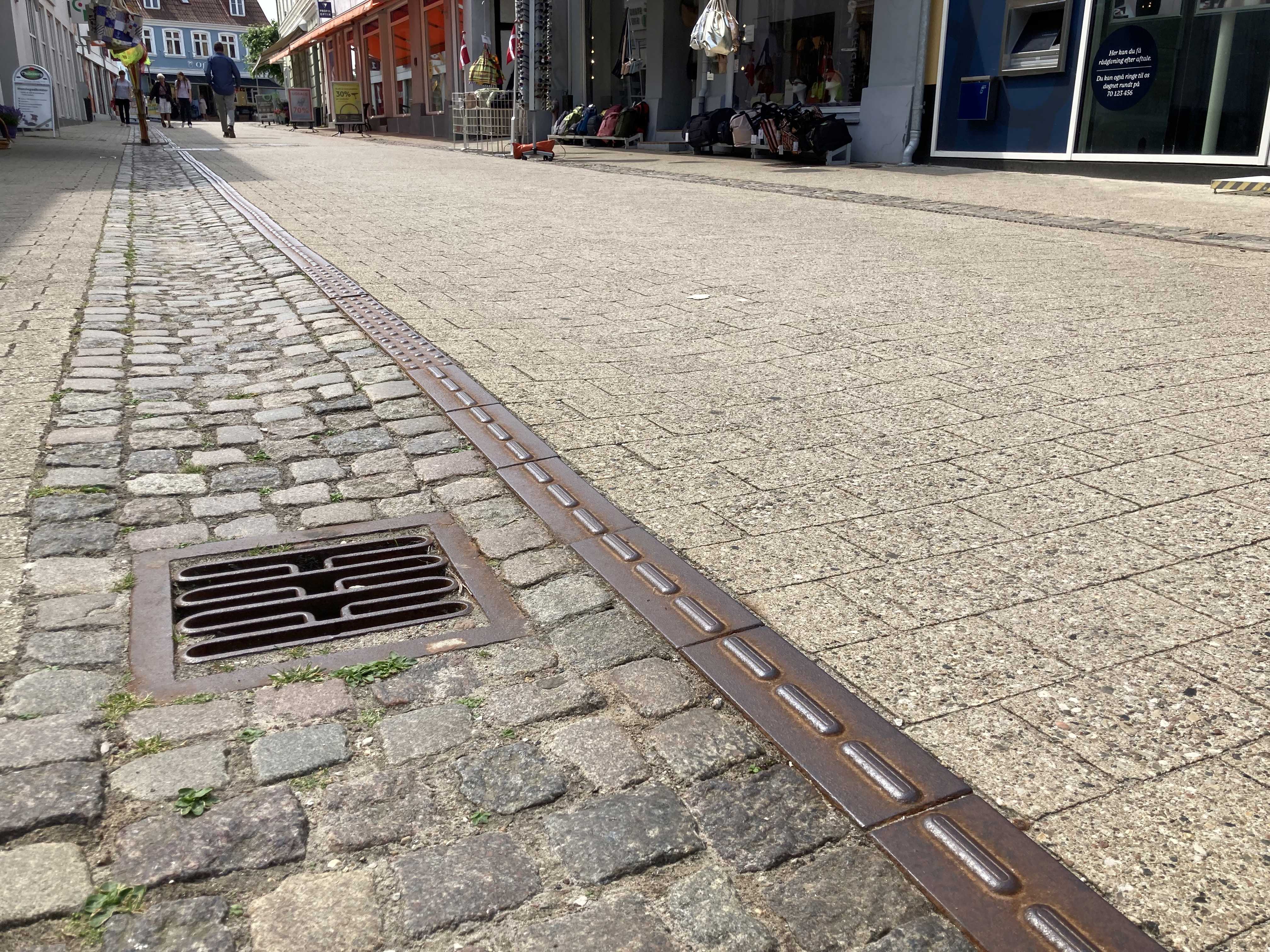 Closeup af brostensbelægningen i Algade, hvor gågaden er i fokus og fremstår nyfejet. I baggrunden fornemmer man handelslivet og butikkerne