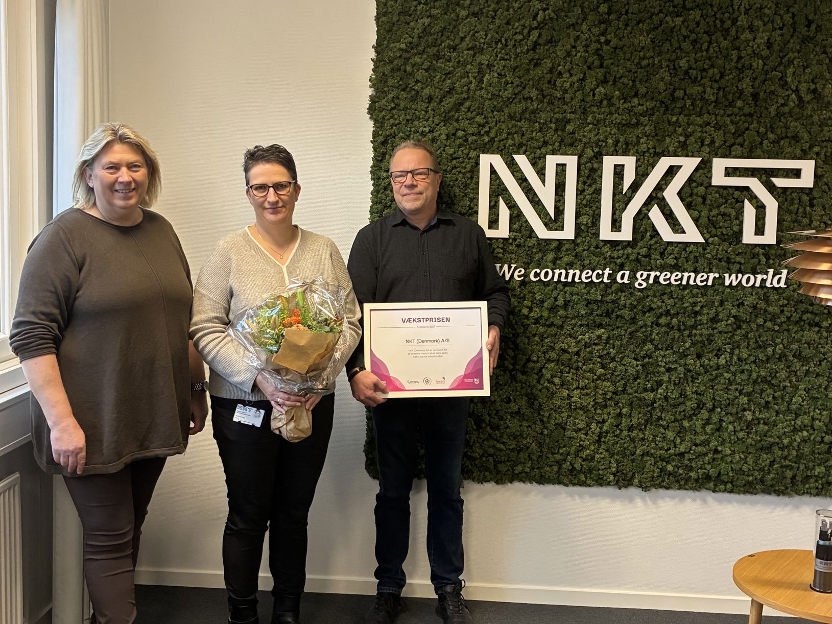 Tre mennesker foran et billedtæppe med teksten NKT