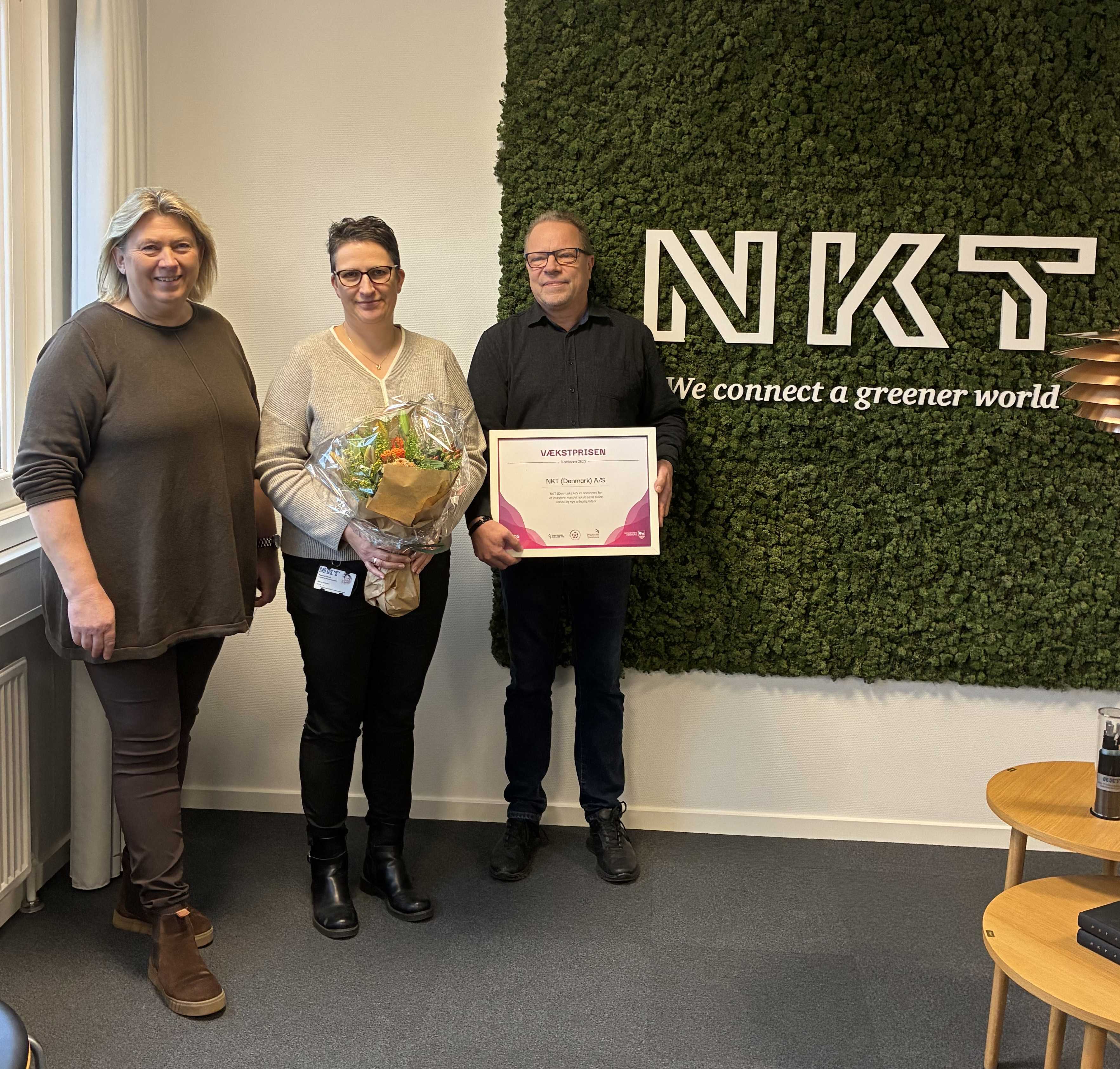 Tre mennesker foran et billedtæppe med teksten NKT