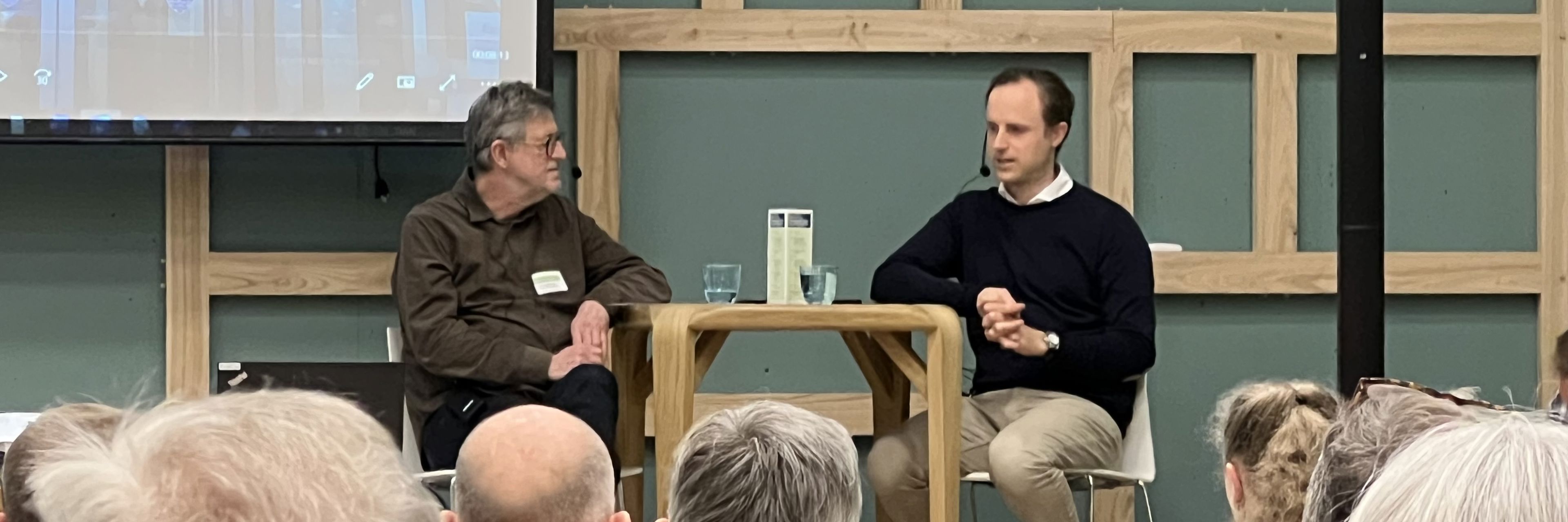 To mænd sidder ved et bord på en scene og taler