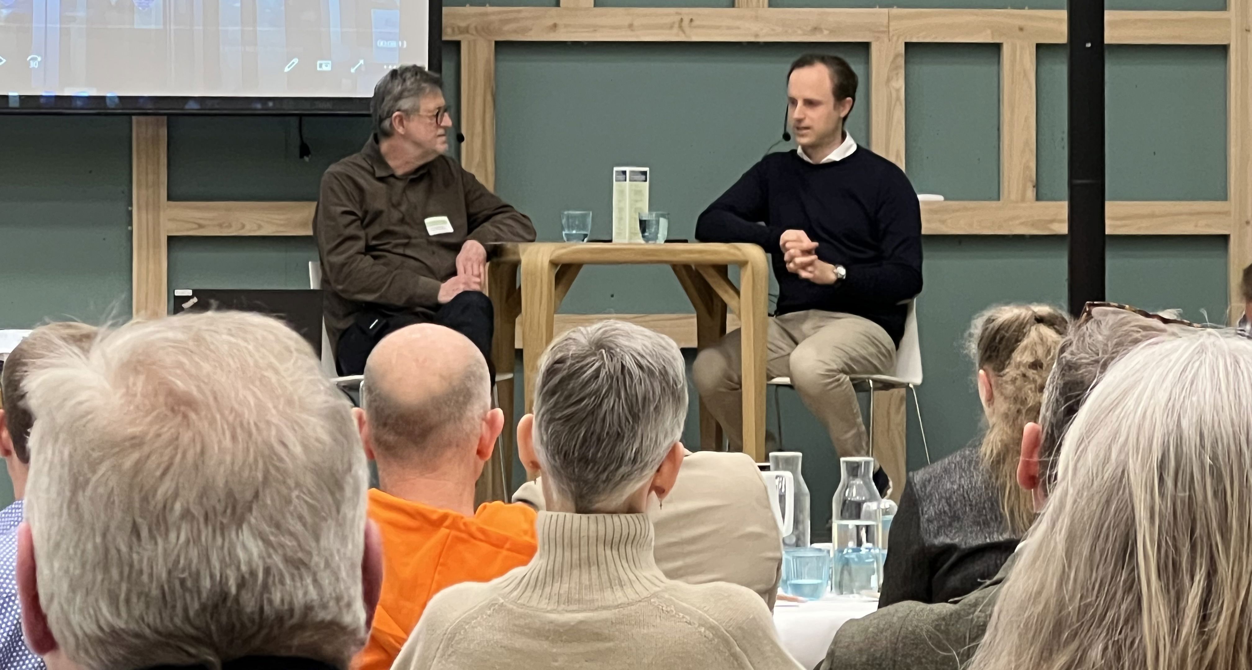 To mænd sidder ved et bord på en scene og taler