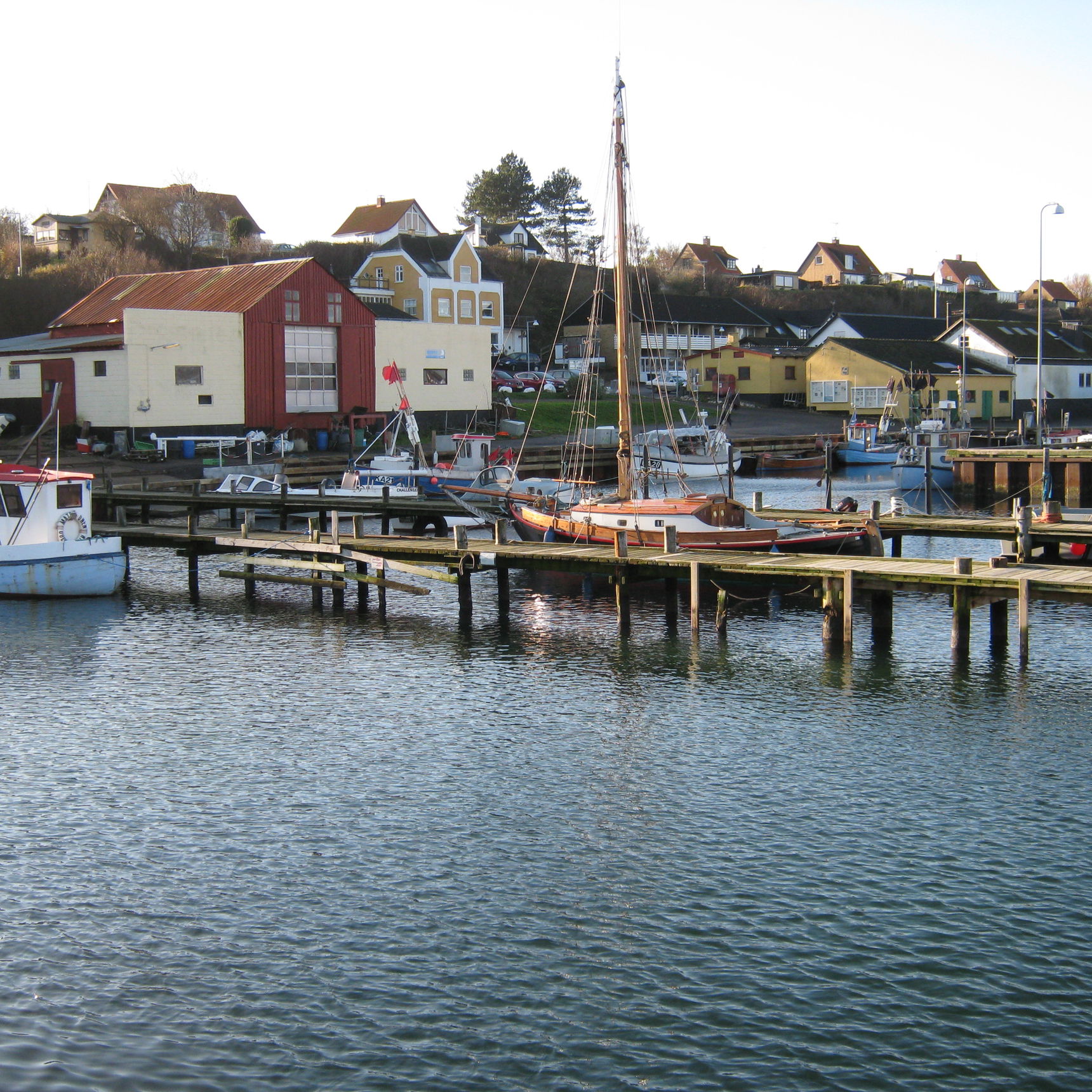Kig fra vandet mod havnefronten. I forgrunden moler og både. I baggrunden huse på havnen.